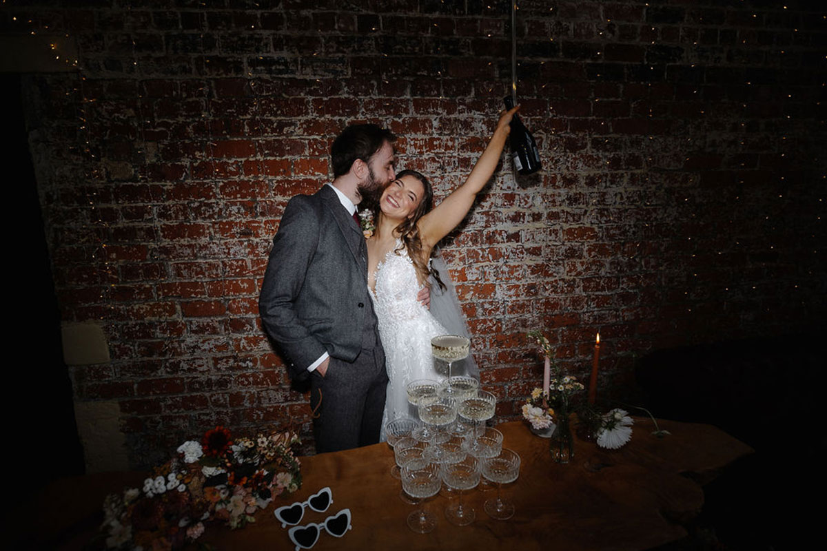 with his arm around her waist, groom kisses bride on the cheek as she smiles widely and lifts a bottle of champagne up in the air, a champagne tower sits on the table in front of them