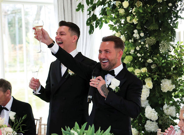 two grooms raise champagne flutes against a green and white foliage background