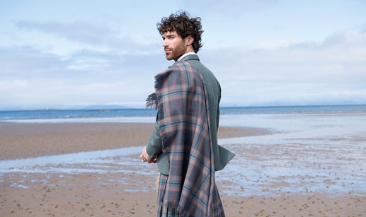 Man with long curled brown hair standing on beach wearing tartan plaid, blue jacket and kilt