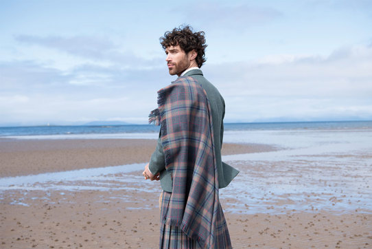 Man with long curled brown hair standing on beach wearing tartan plaid, blue jacket and kilt