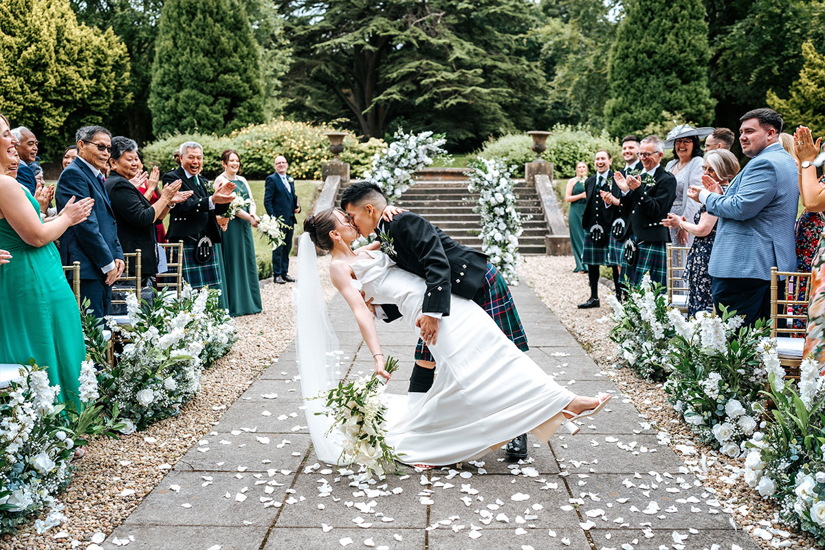 A bride and groom dip and kiss in the middle of an outdoor aisle while their guests look from their seats and clap