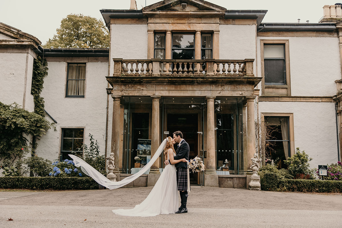 Bride and groom sharing a kiss outside a white manor house with columns and a long flowing veil