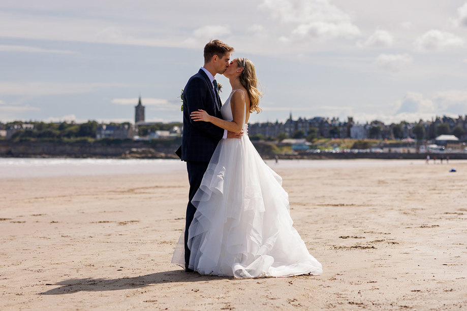 A couple sharing a kiss on a sandy beach, clothed in wedding attire, with the Old Course Hotel and cloudy blue sky in the background