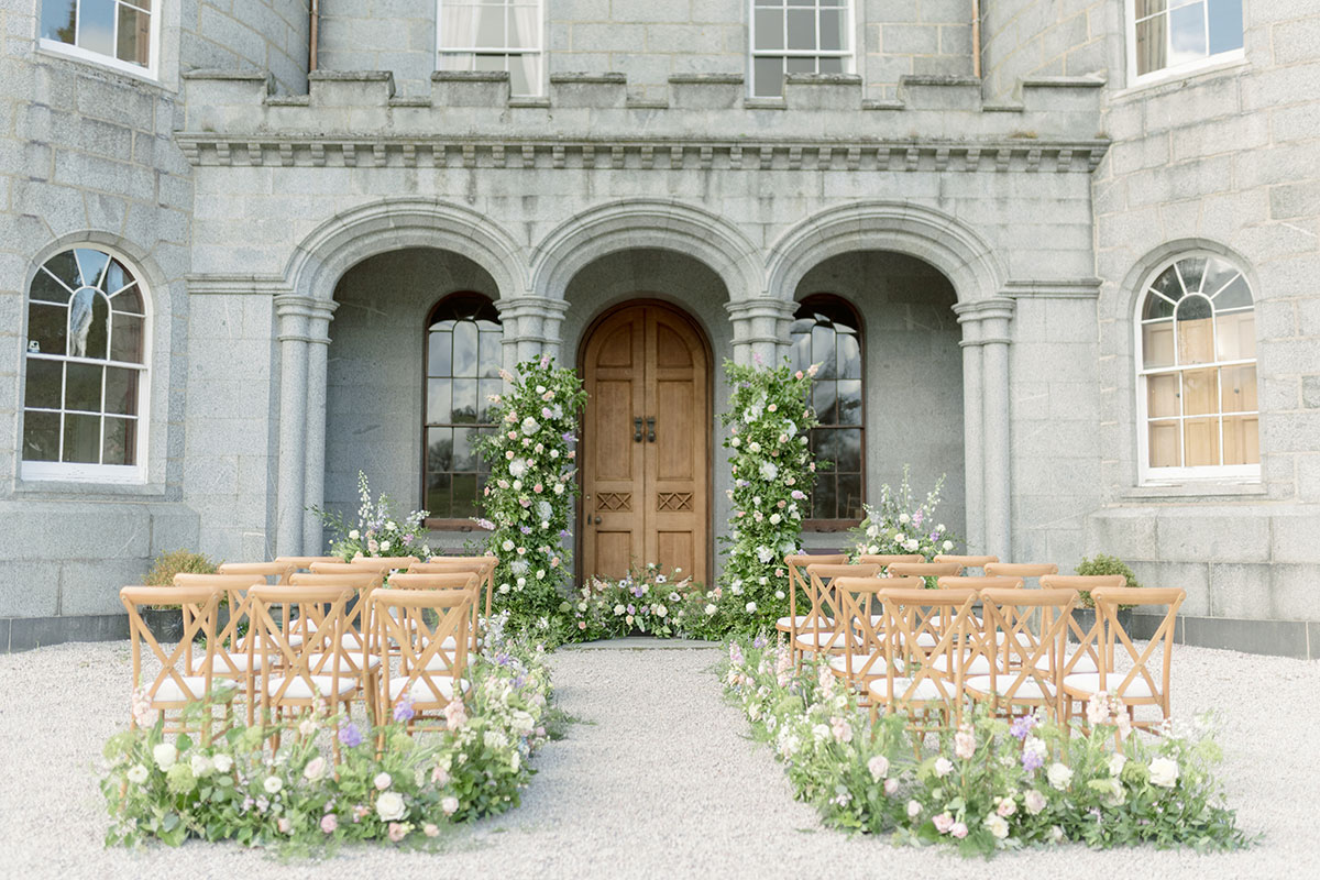Outdoor wedding ceremony setup at Cluny Castle with floral-lined aisle, wooden cross-back chairs and the castle entrance as a backdrop