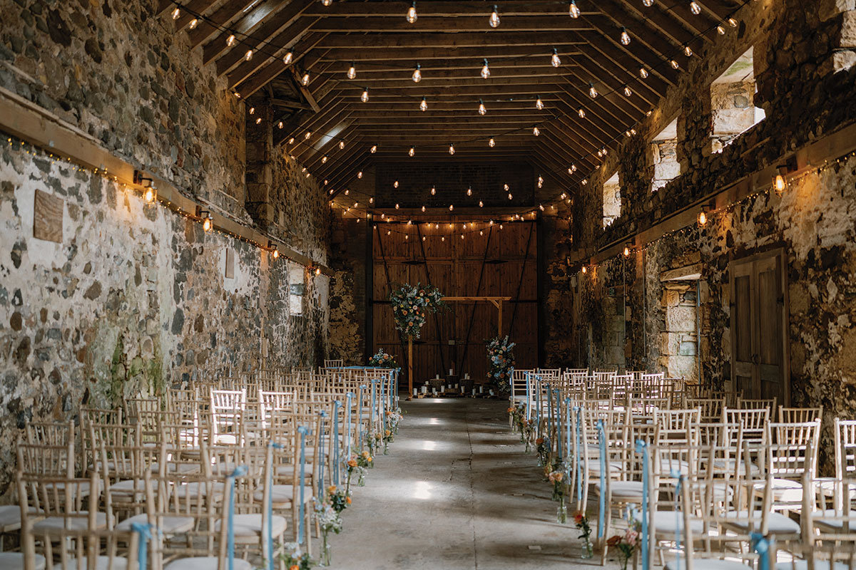 Rustic wedding ceremony set-up inside Pratis Barns in Fife with wooden chairs, floral aisle decorations and festoon lighting