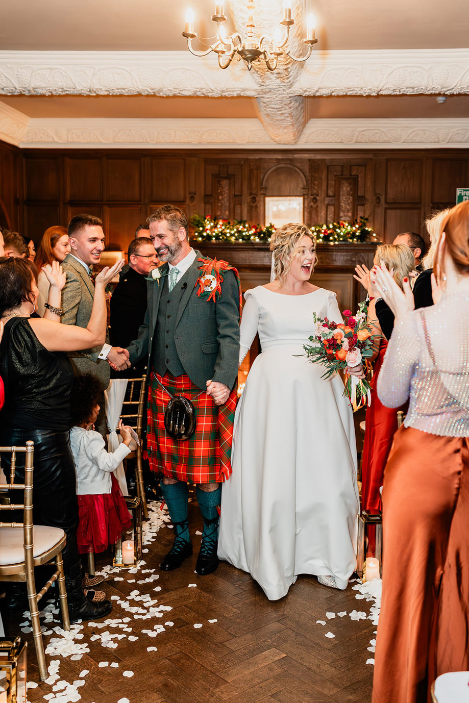 bride in white wedding dress and veil and groom in red and green tartan kilt outfit walk hand in hand up the aisle smiling and looking to either side at their friends and family