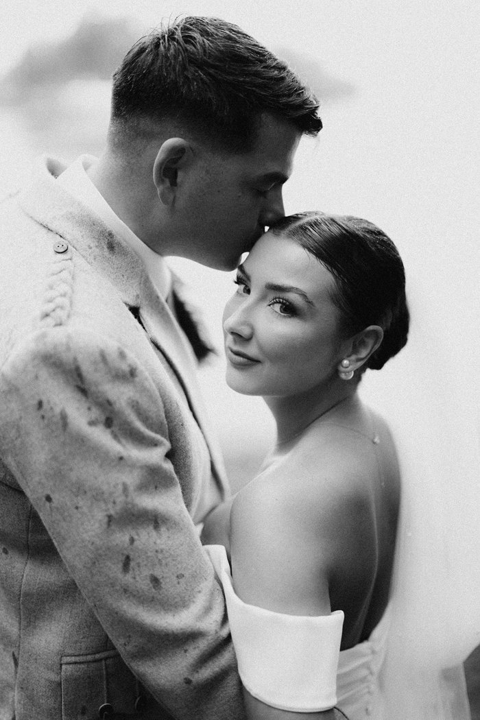 black and white image of bride in strapless wedding dress with off the shoulder straps and long veil being held in arms of tall groom who kisses her softly on the forehead