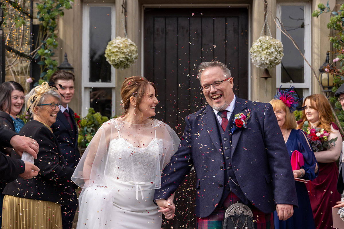 A bride and groom walk out of a building holding hands as their guests throw confetti over them