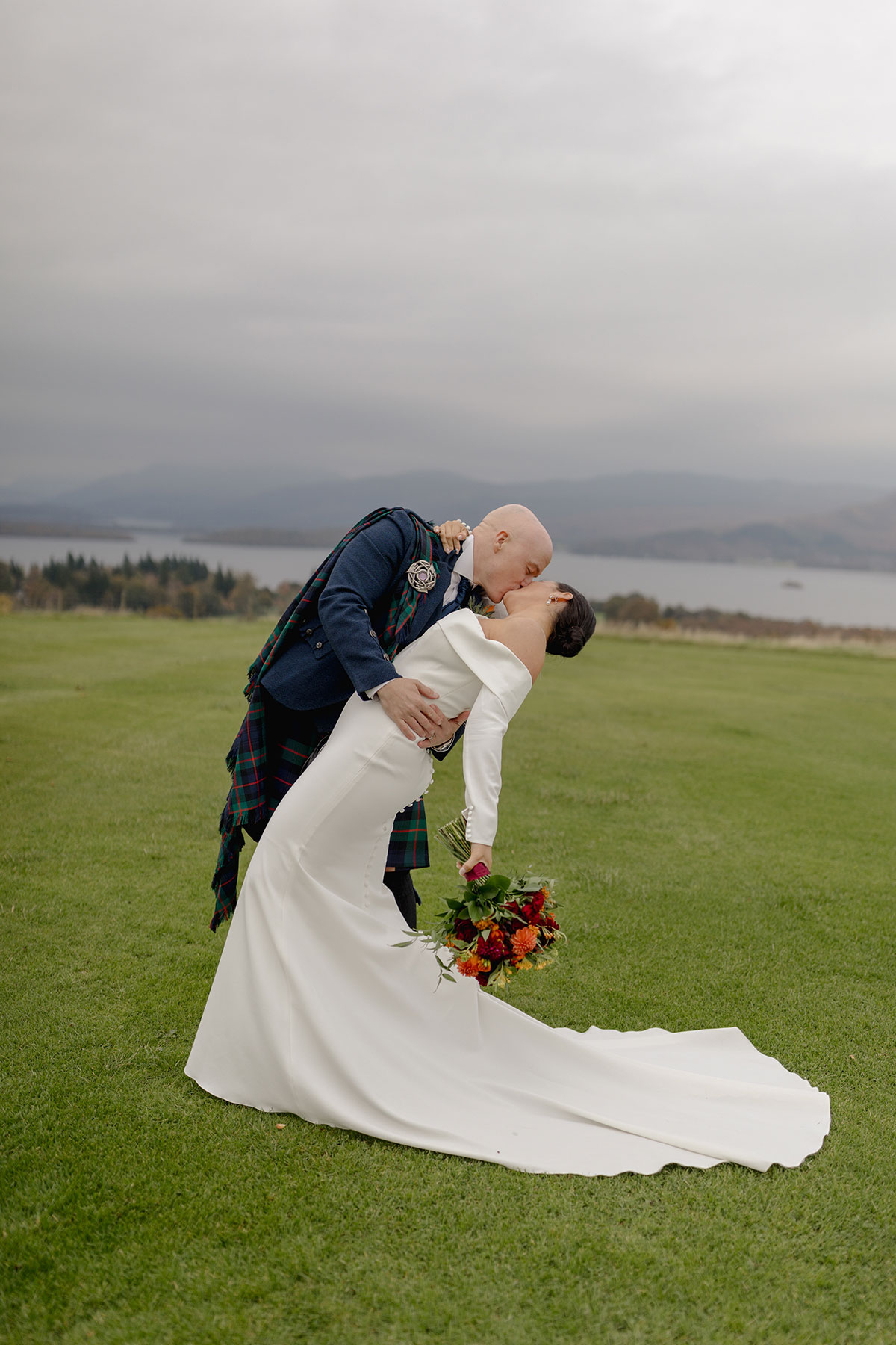 Newlyweds kiss on the lawn at Ardoch Loch Lomond, with the bride holding a colourful bouquet and Loch Lomond and the surrounding hills in the background.