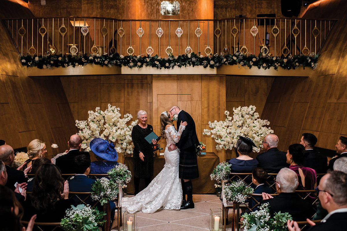 Wedding ceremony in Fingal’s Ballroom with floral arrangements, wooden interiors and guests watching the couple’s first kiss