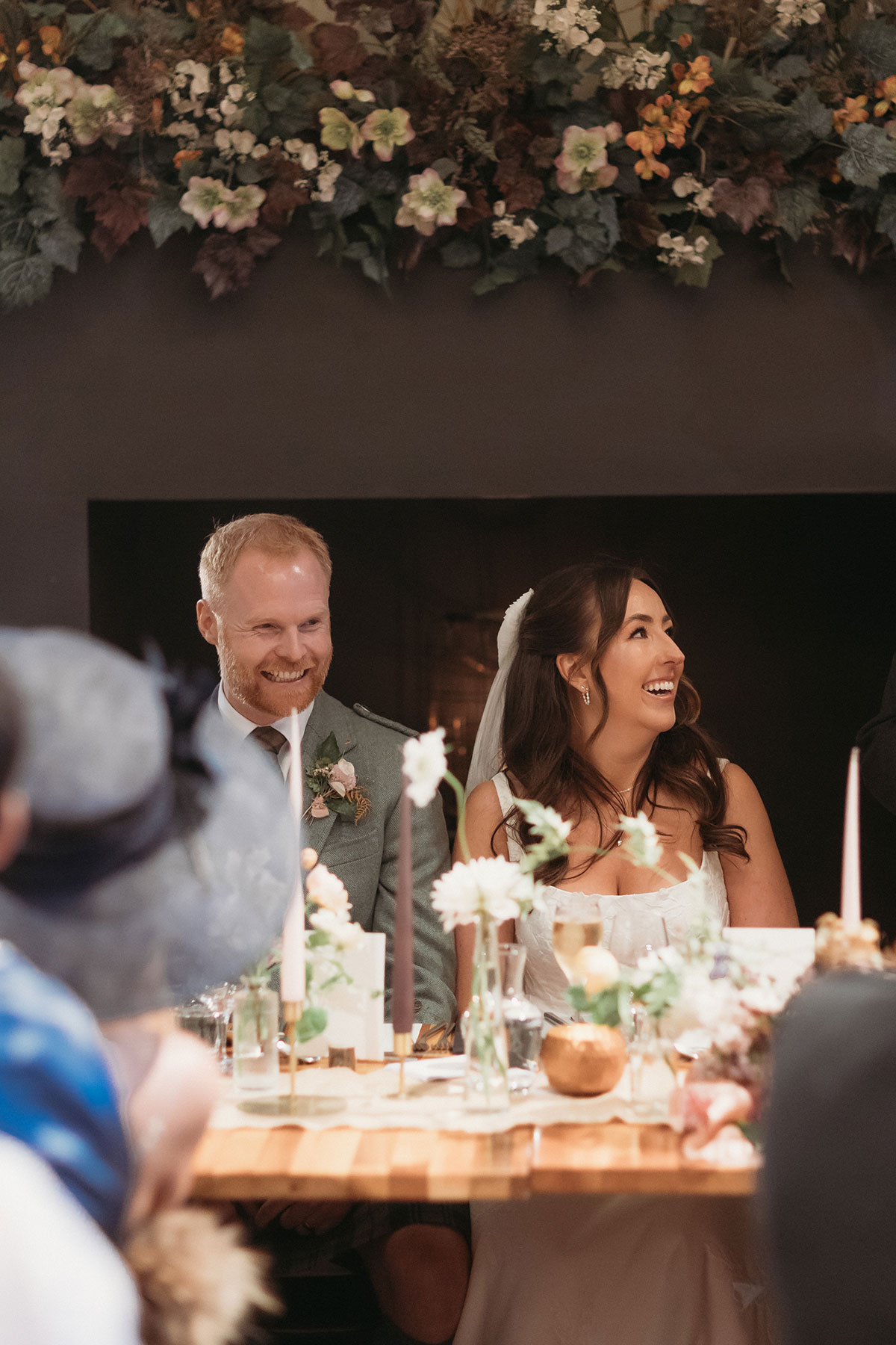a laughing bride and groom seated at a table decorated with flowers and tall candles.