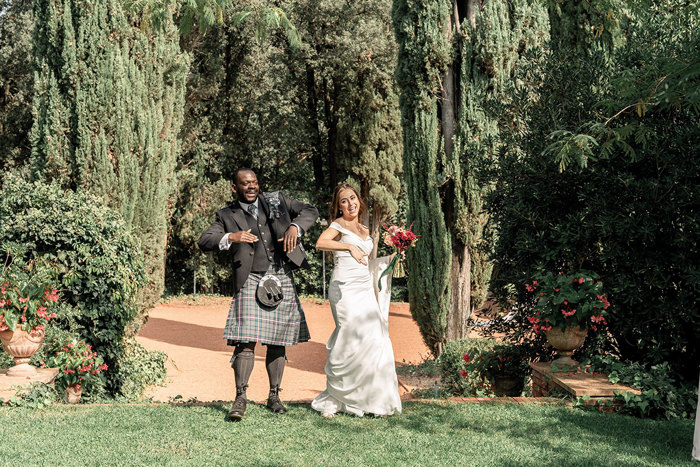 a bride in an off the shoulder gown and a groom in a dark tartan kilt outfit seemingly dance across a grassy area