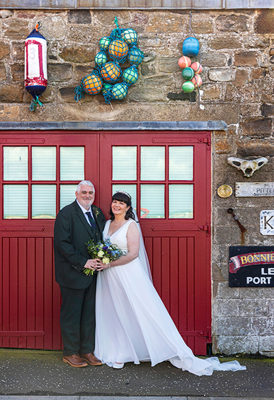 a bride and groom standing in front of the red doors of a boat shed decorated with sea paraphernalia