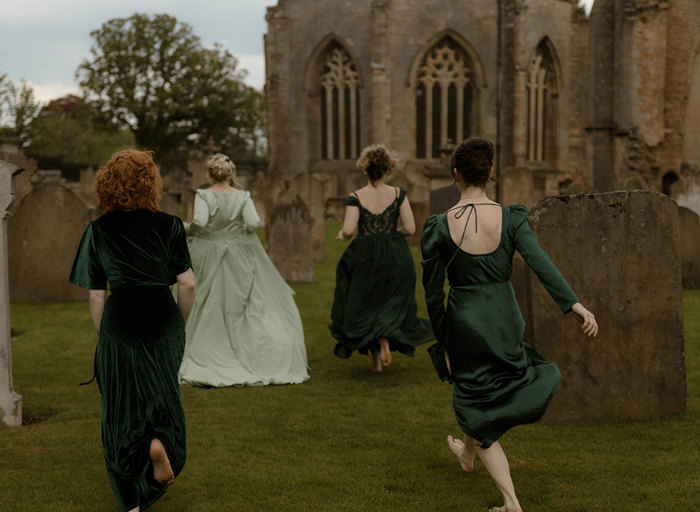 a bride wearing a pale green dress and three bridesmaids wearing dark green dresses running through a graveyard at Elgin Cathedral