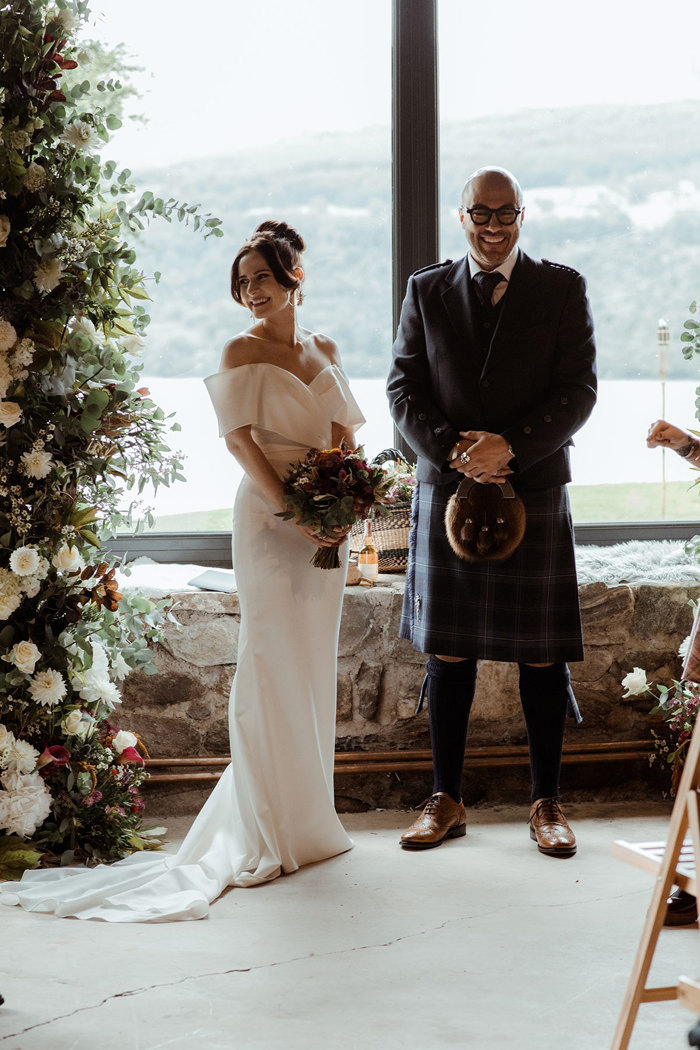 groom in blue, grey and white tartan kilt outfit stands in front of large scenic window with bride to his side in off the shoulder white wedding dress, holding a bouquet of flowers in front of her waist