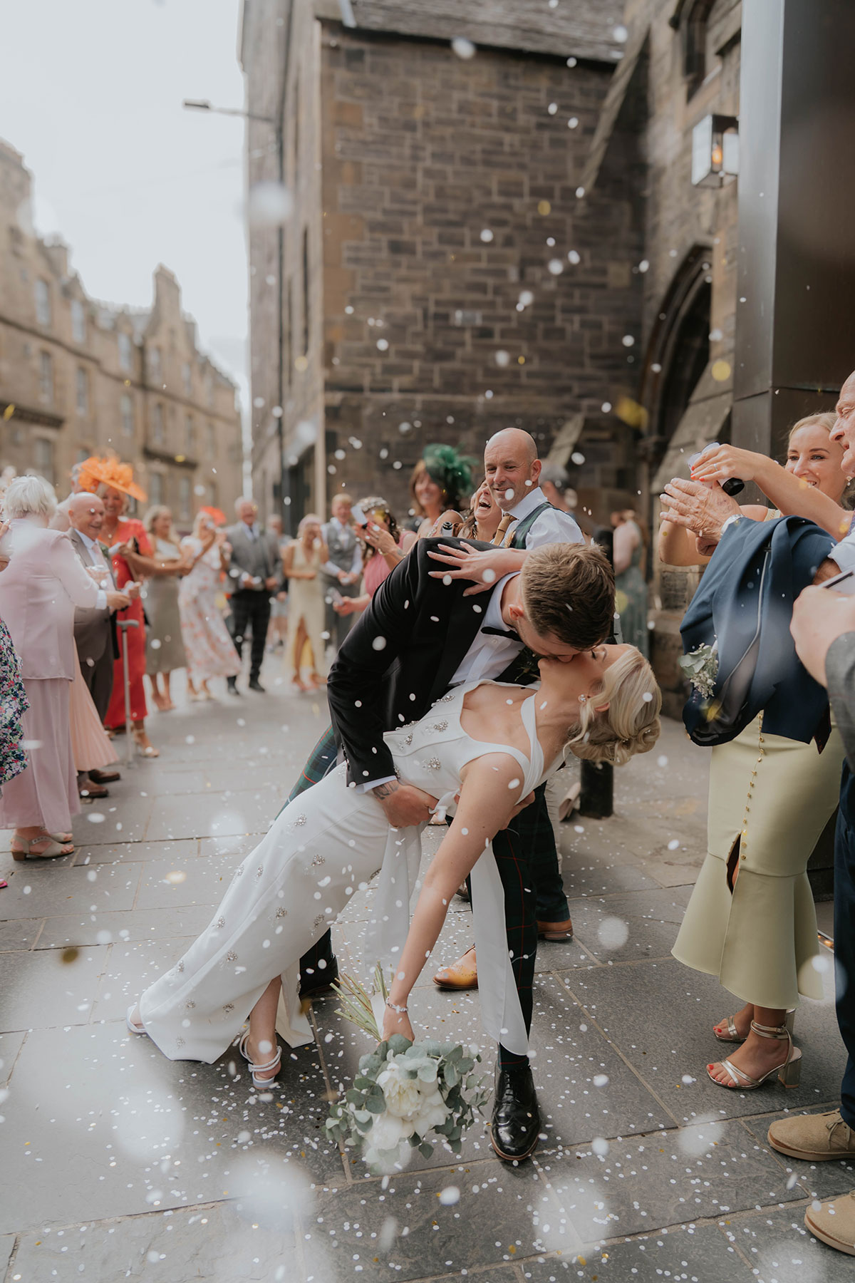 Groom dipping and kissing the bride as guests throw confetti outside their Edinburgh wedding ceremony