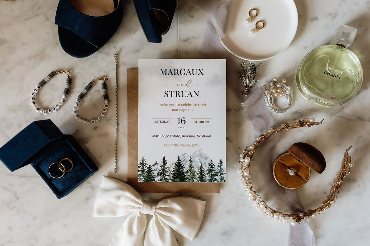 Wedding invitation flat lay with rings, bridal accessories, perfume and jewellery for a Scottish wedding at Mar Lodge Estate