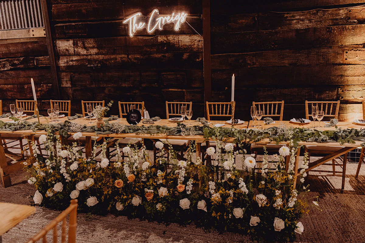 A long wooden wedding table decorated with greenery and candles, with a line of fresh florals arranged along the front.