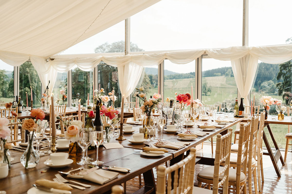 A wedding reception inside a marquee with clear sides, wooden tables, floral décor and views across the estate.