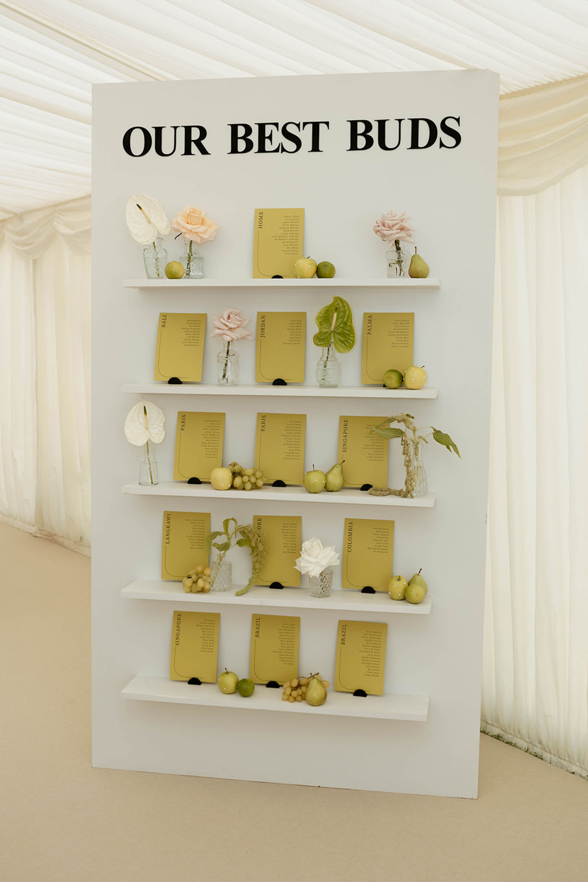 Wedding seating plan display with yellow cards, fresh fruit and floral styling on white shelves.