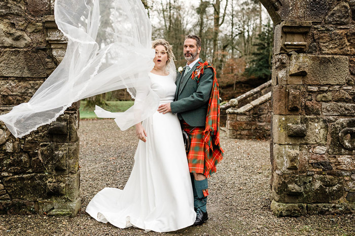 bride in white wedding dress and veil and groom in red and green tartan kilt outfit face each other under an outdoor stone arch as bride's veil blows in the wind