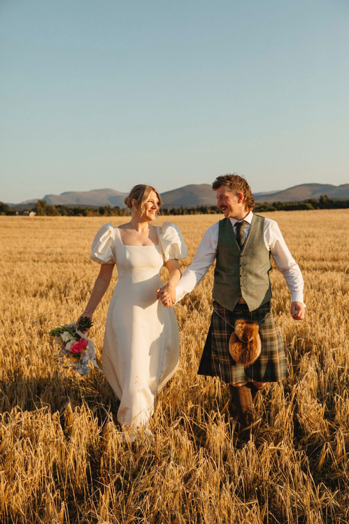 Bride and groom holding hands in wheat field during sunset portraits at Cairngorms Highlands wedding