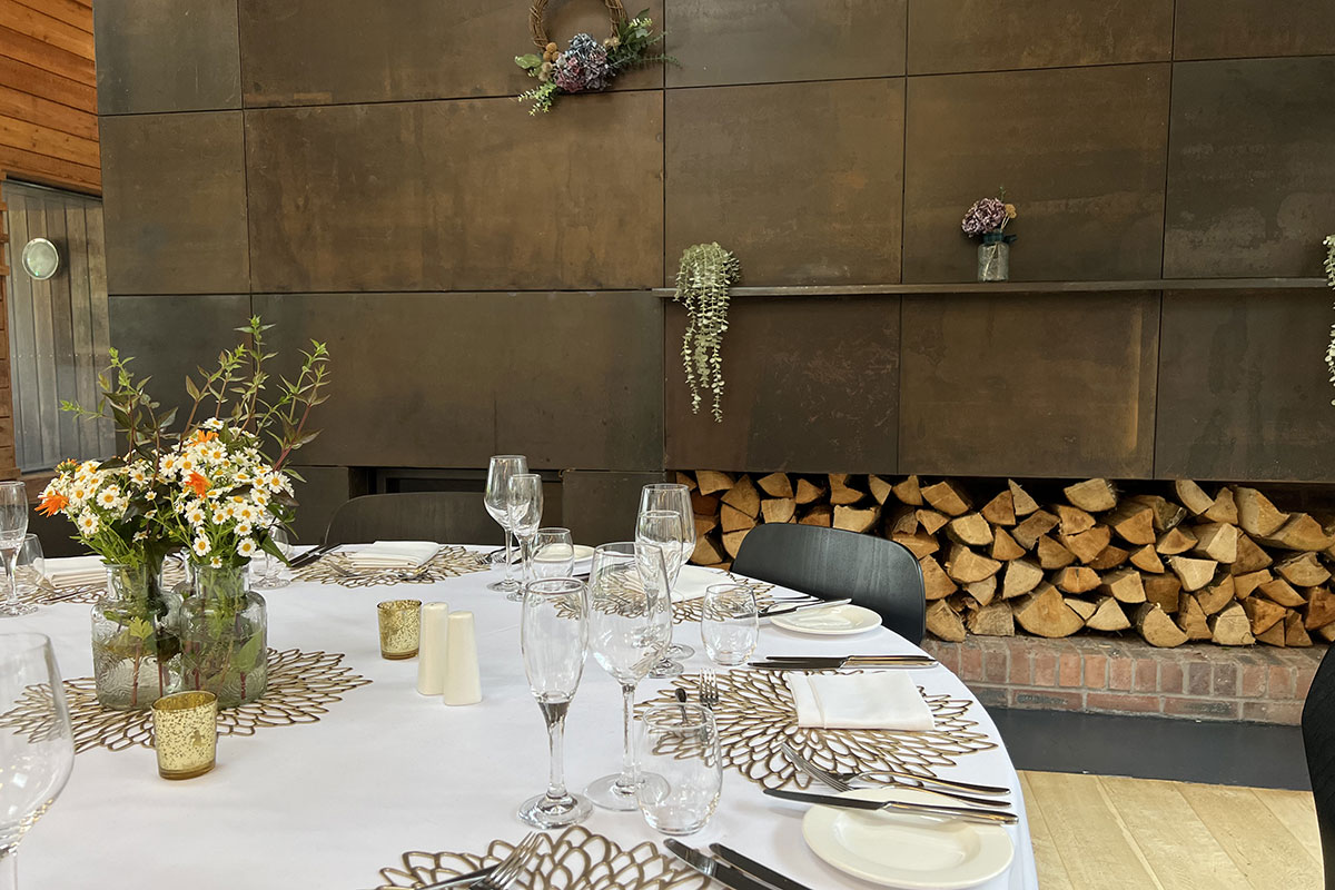 view of the top of a round dining table set with glassware, golden flower placemats, flowers in the middle and logs of firewood piled in the background in the wall