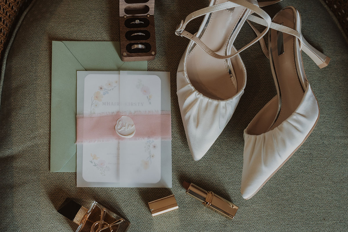 Bridal flat lay with ivory wedding heels, invitations, perfume and rings on textured fabric background