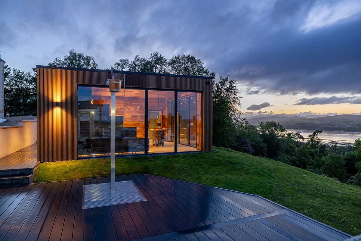 Glass-fronted building glowing with warm light at dusk, surrounded by trees and overlooking a calm body of water