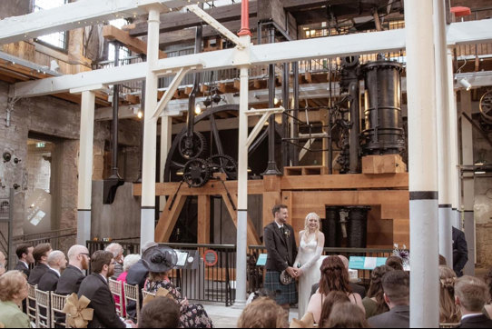a bride and groom standing in front of the old jute mill at Verdant Works in Dundee during a wedding ceremony as guests seated in rows look on 
