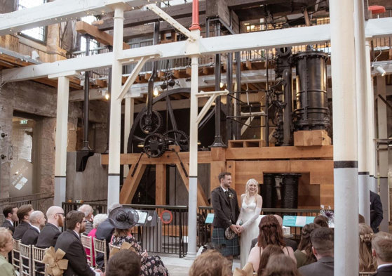 a bride and groom standing in front of the old jute mill at Verdant Works in Dundee during a wedding ceremony as guests seated in rows look on 