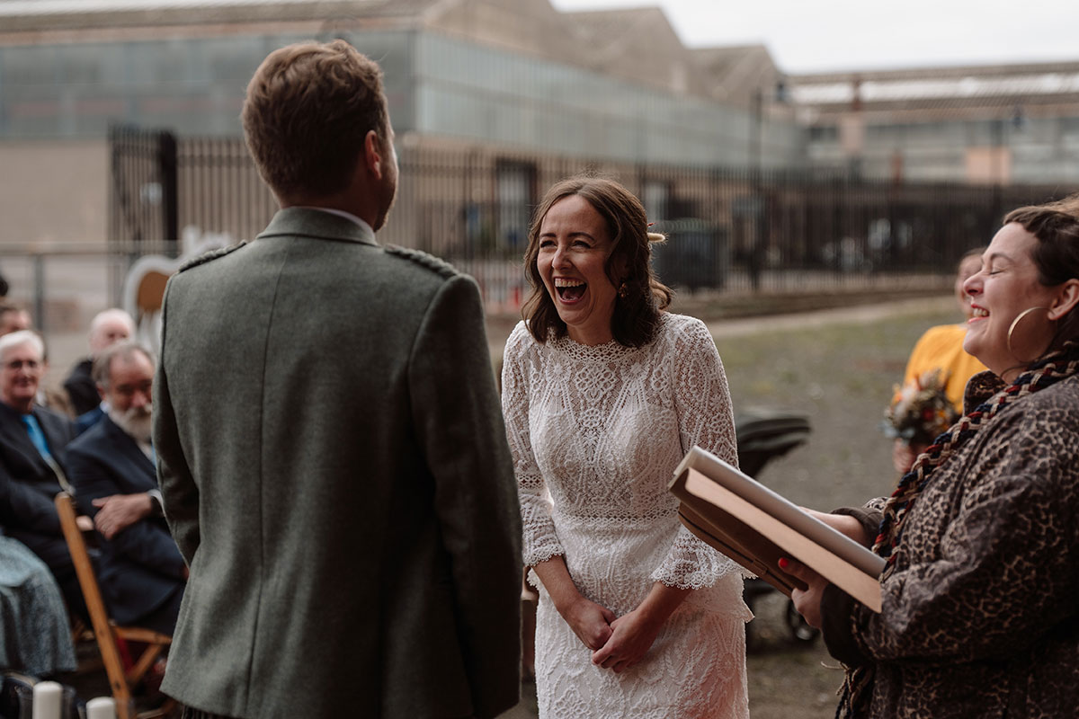 Bride laughing during an outdoor humanist wedding ceremony, standing opposite the groom while guests watch from their seats