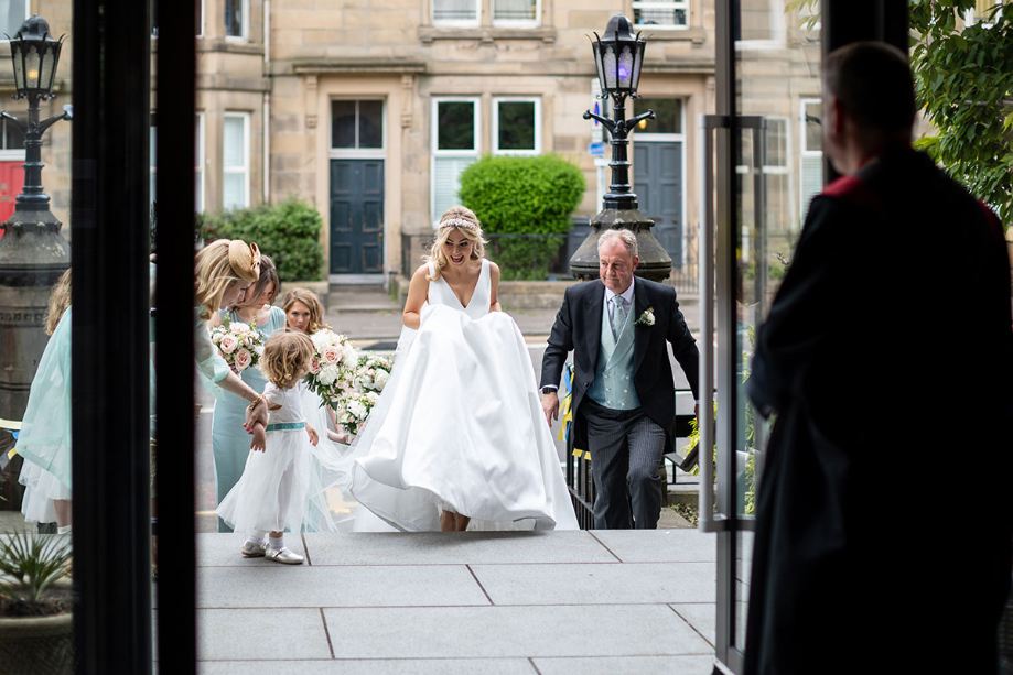 Bride walks into wedding venue with bridesmaids waiting at the side and father behind her