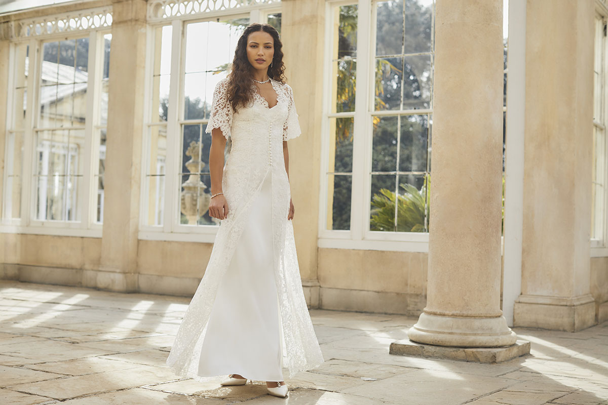 curly haired brunette woman stands in empty tiled room with large column wearing slinky white dress with short-sleeved lace overcoat