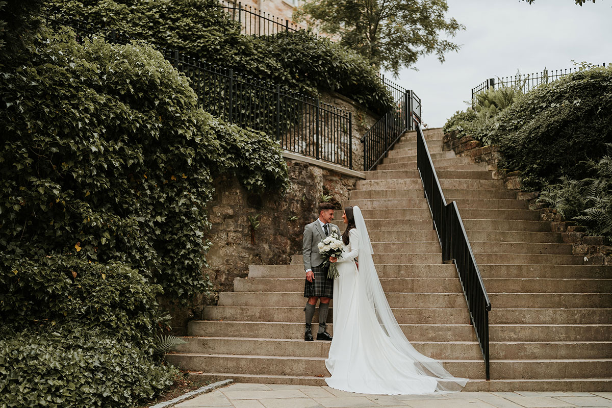bride and groom smile at one another while standing on steps at crossbasket castle on wedding day