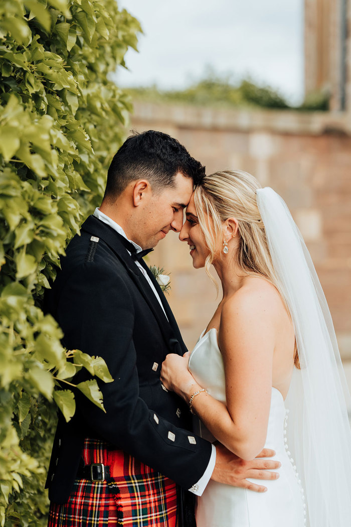 A groom in a red kilt and a bride in wedding attire standing with their foreheads pressed together.