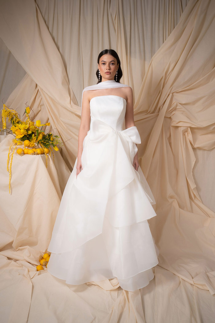 bride with dark brown hair wears strapless ballgown wedding dress with a bridal scarf drapped back over her shoulders and a ruffled bow on the front of her dress as she stands in front of a dark cream sheet backdrop also decorated with an assortment of yellow flowers and lemons off to one side