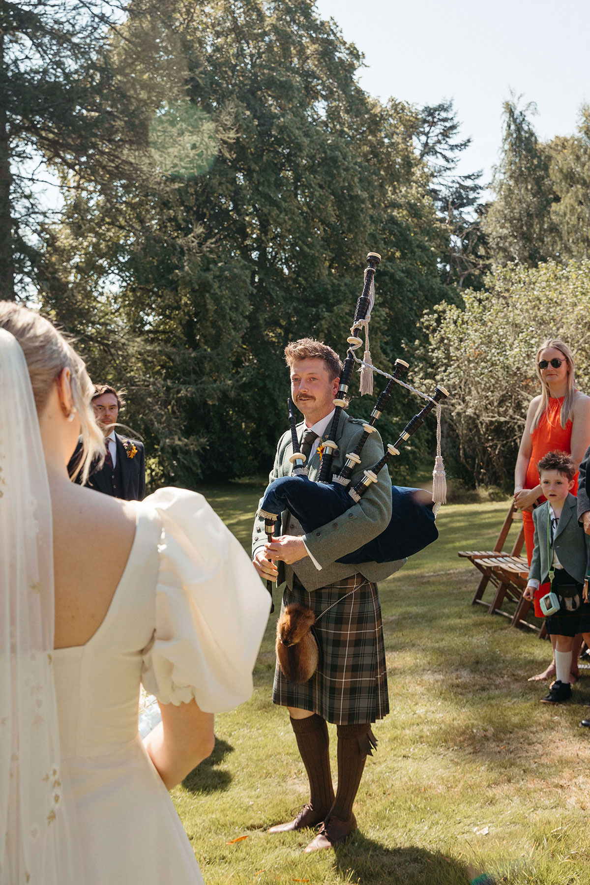 Groom plays bagpipes during outdoor Highland wedding ceremony at bride’s family home in Nethy Bridge