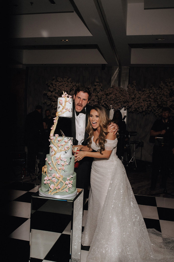 a bride and groom cutting a pale green four-tier cake decorated with pink flowers and a dog figurine. They have a shocked expression on their faces.