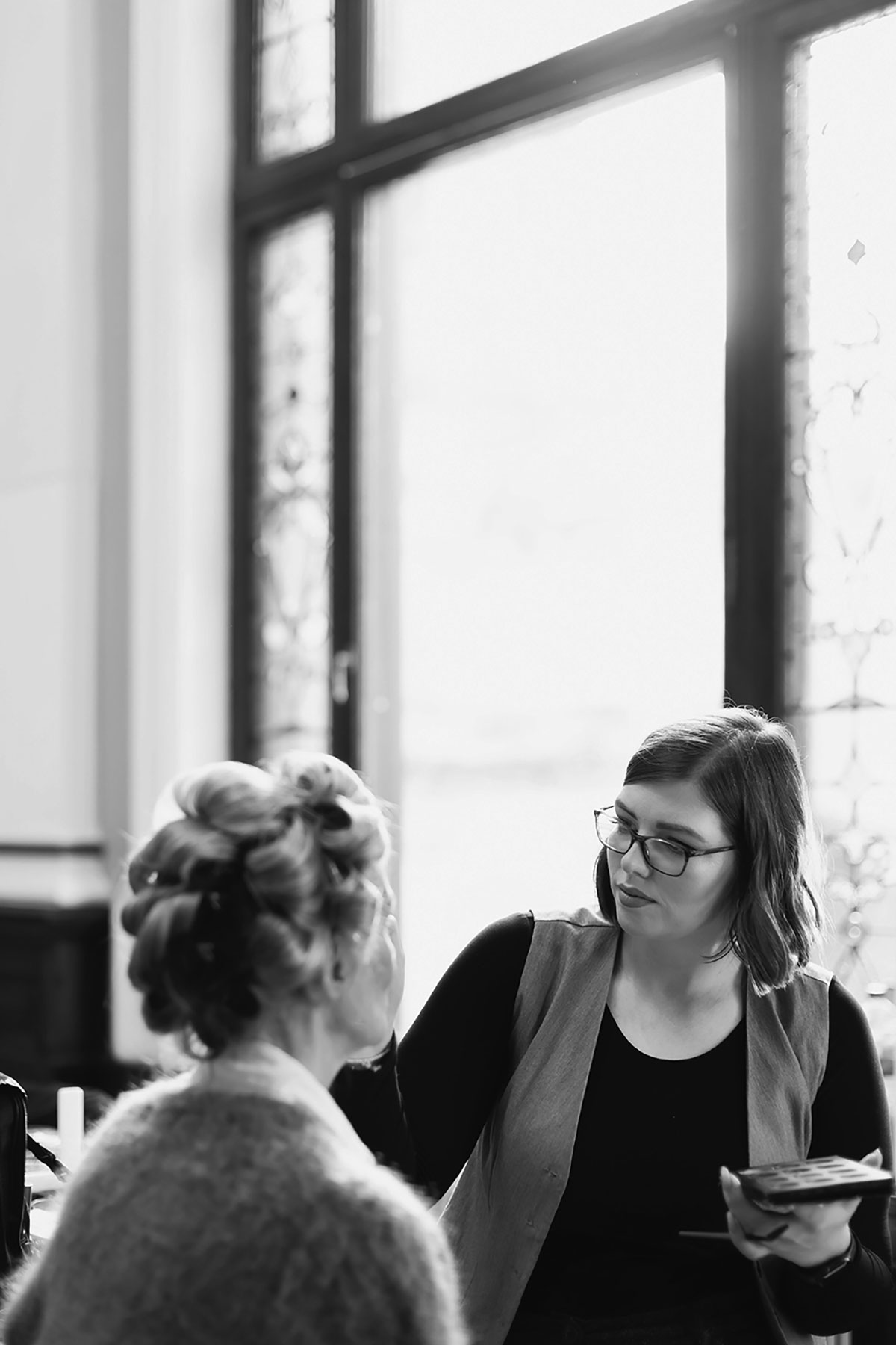 Black-and-white candid of makeup artist applying bridal makeup beside stained-glass window