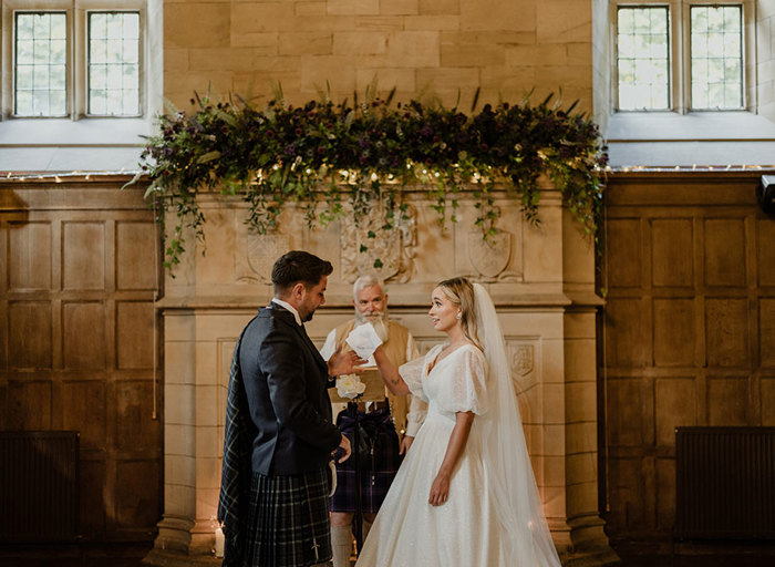 a bride passes a groom an embroidered handkerchief as they stand in front of an officiant and a large stone fireplace that's draped with greenery, foliage and fairy lights