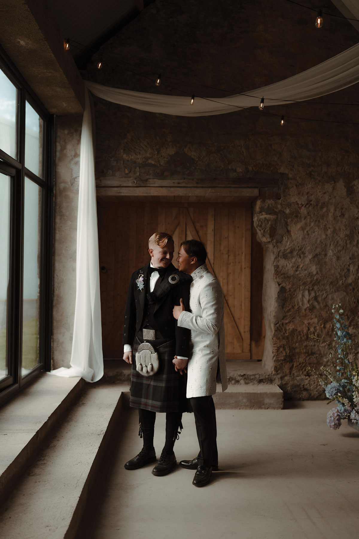 Same-sex newlyweds posing inside rustic stone barn wedding venue near Falkirk, Scotland