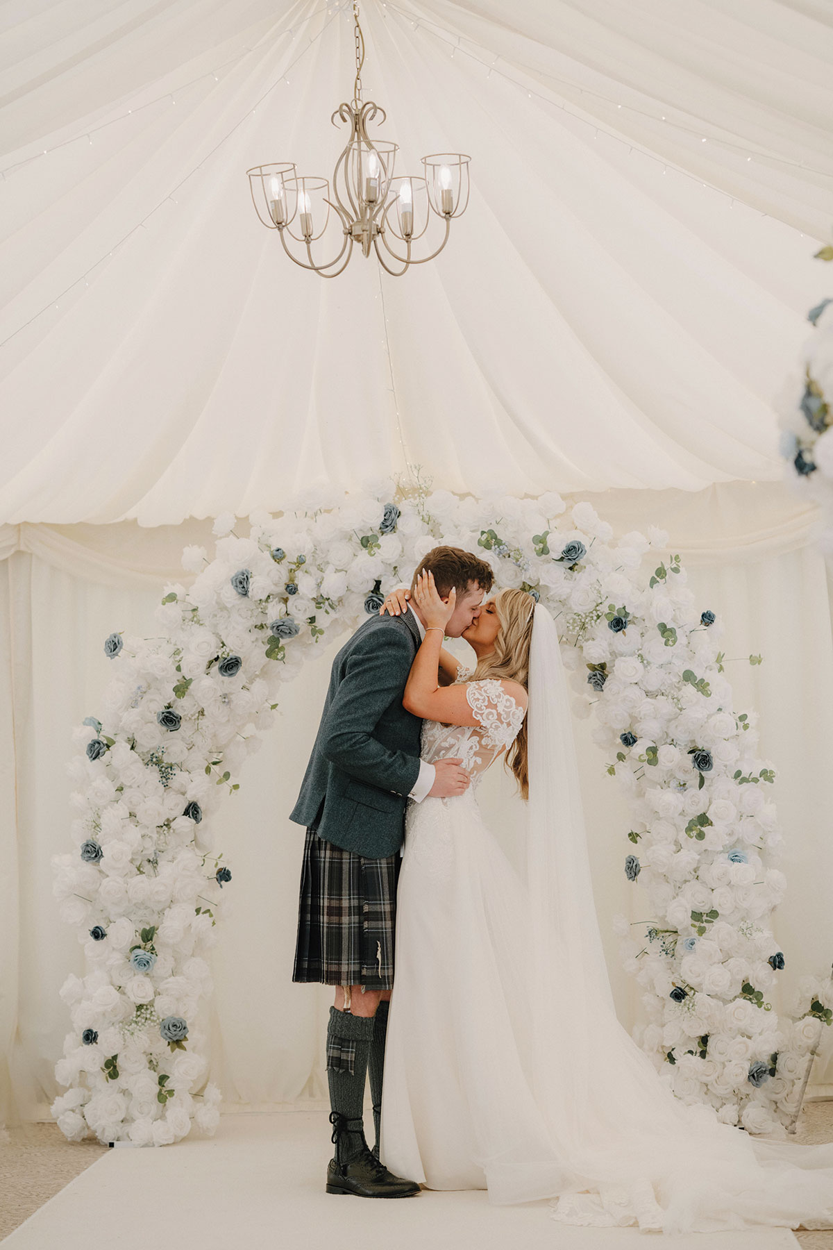 bride and groom kiss for the first time as married couple inside marquee at cornhill castle that's decorated with a white and blue floral arch