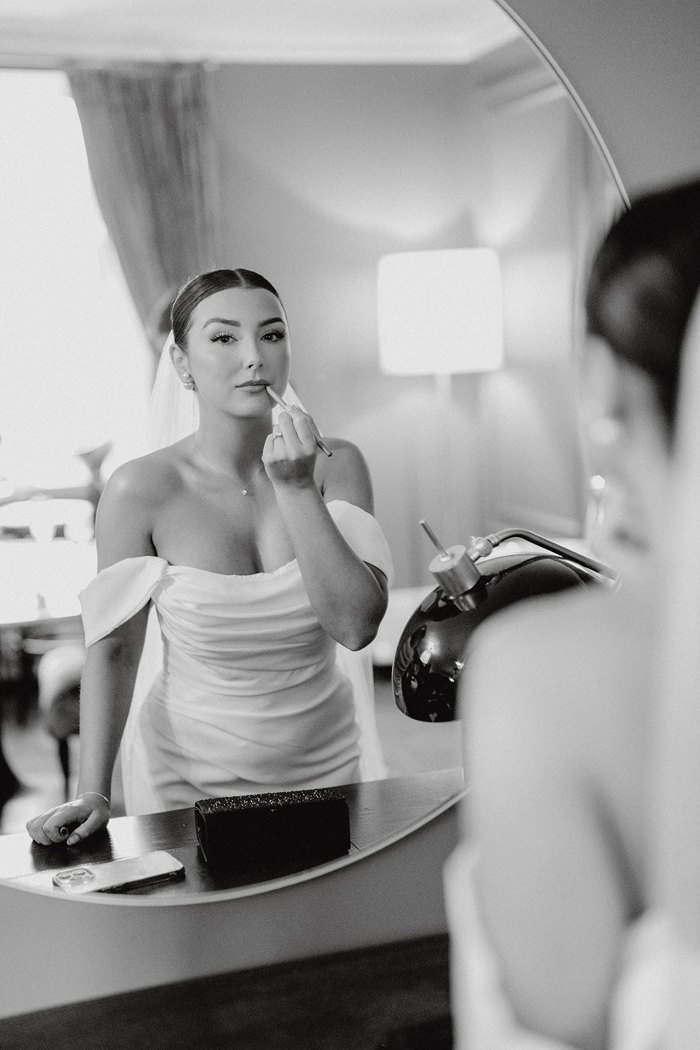 black and white image of bride in strapless wedding dress with off the shoulder straps applying the finishing touches to her makeup in the mirror