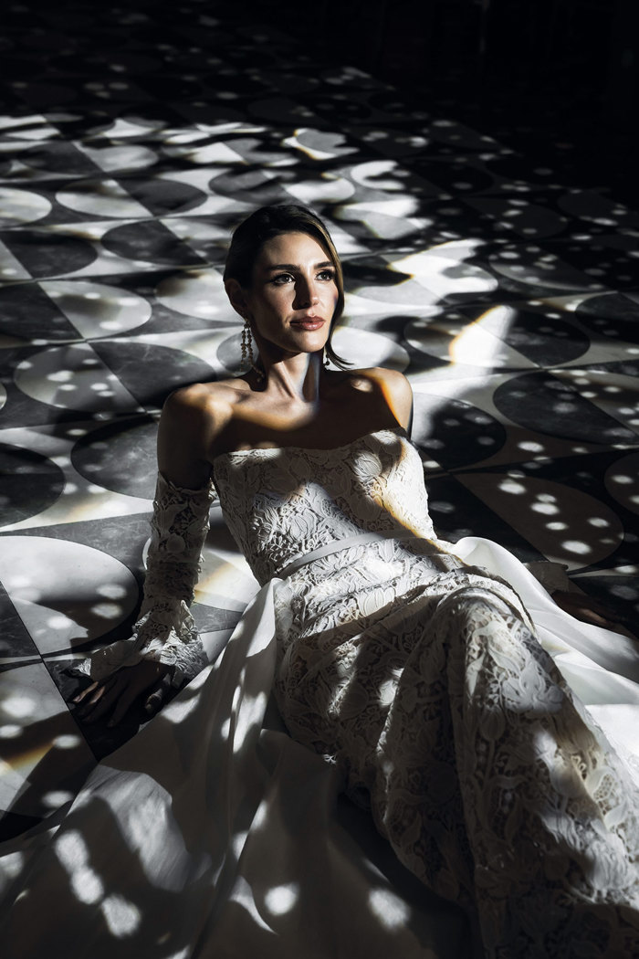 a brunette bride wearing a lace wedding dress from Eleganza Sposa sitting on a black and white circular-patterned floor below dappled lighting at the Exchange wedding venue in Glasgow