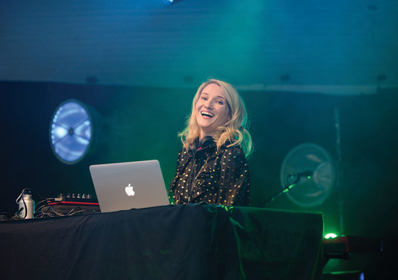 Blonde smiling DJ with apple macbook against a black backdrop