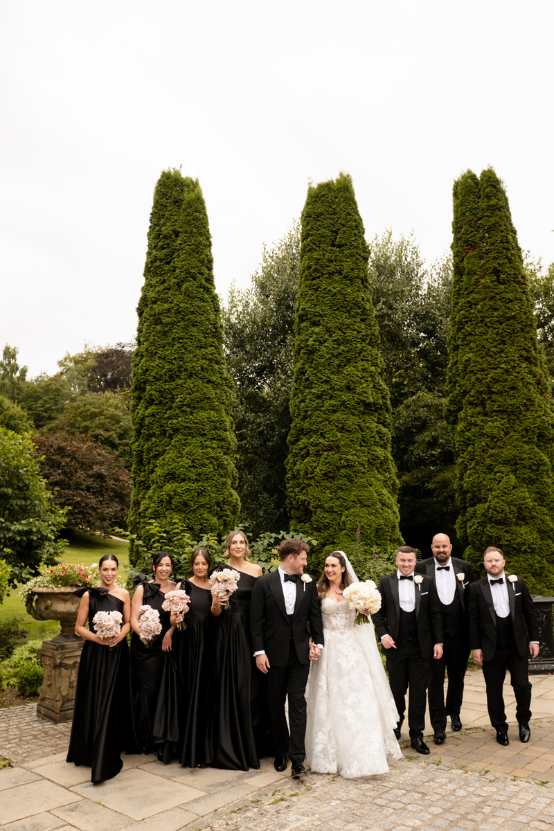 Bride and groom with bridesmaids in black dresses and groomsmen in black tie in the gardens at Crossbasket Castle.
