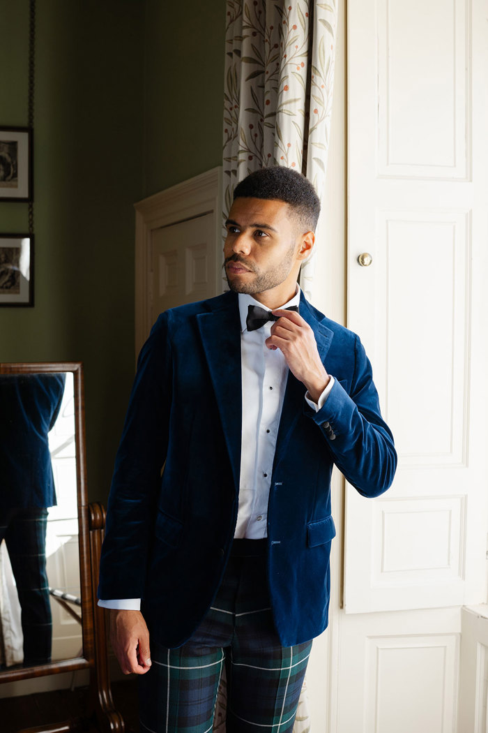 A man wearing green tartan trousers and a navy velvet suit jacket adjusts his bowtie