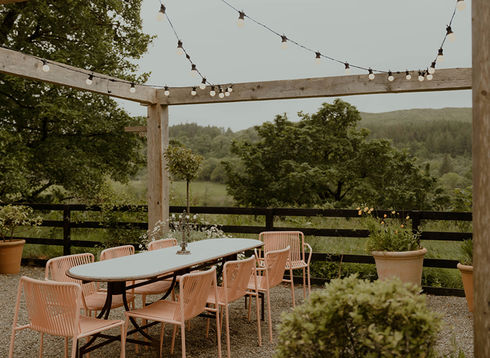 a long table surrounded by peach garden seating under a fairy light canopy and wooden frame amid a lush garden setting