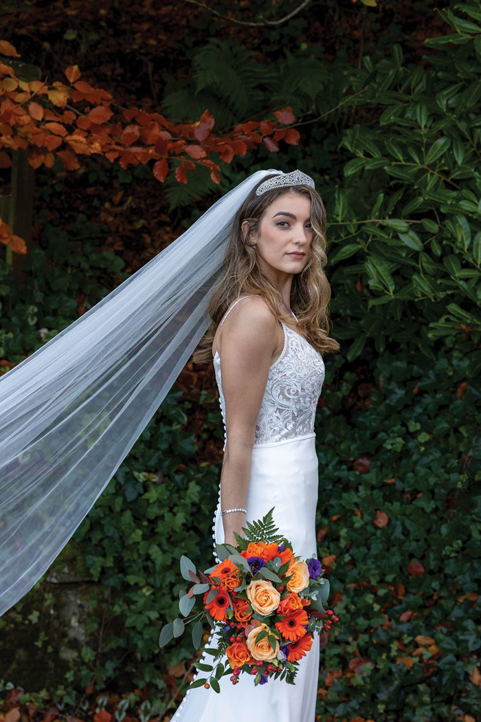 bride stands with wedding dress and long veil on while holding autumnal flowers